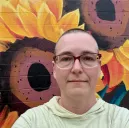 a short haired person wearing glasses and a yellow shirt, in front of a colorful mural with sunflowers
