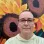a short haired person wearing glasses and a yellow shirt, in front of a colorful mural with sunflowers