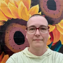 a short haired person wearing glasses and a yellow shirt, in front of a colorful mural with sunflowers